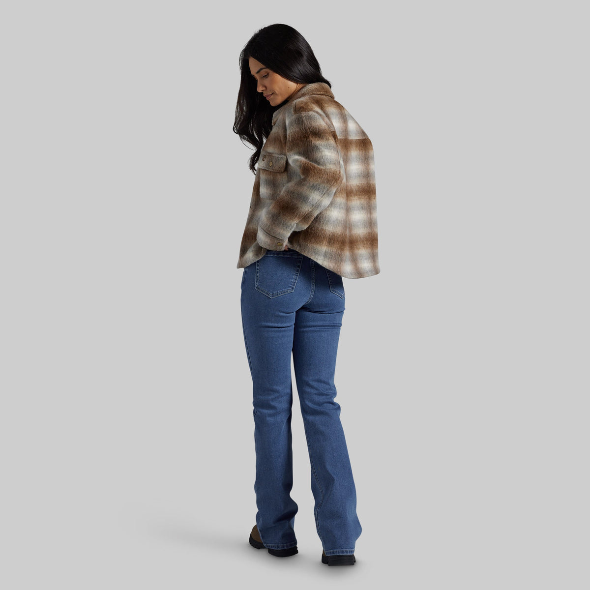 A woman wearing a brown and beige plaid cropped flannel jacket and blue jeans, shown from a side-back angle against a solid gray background.