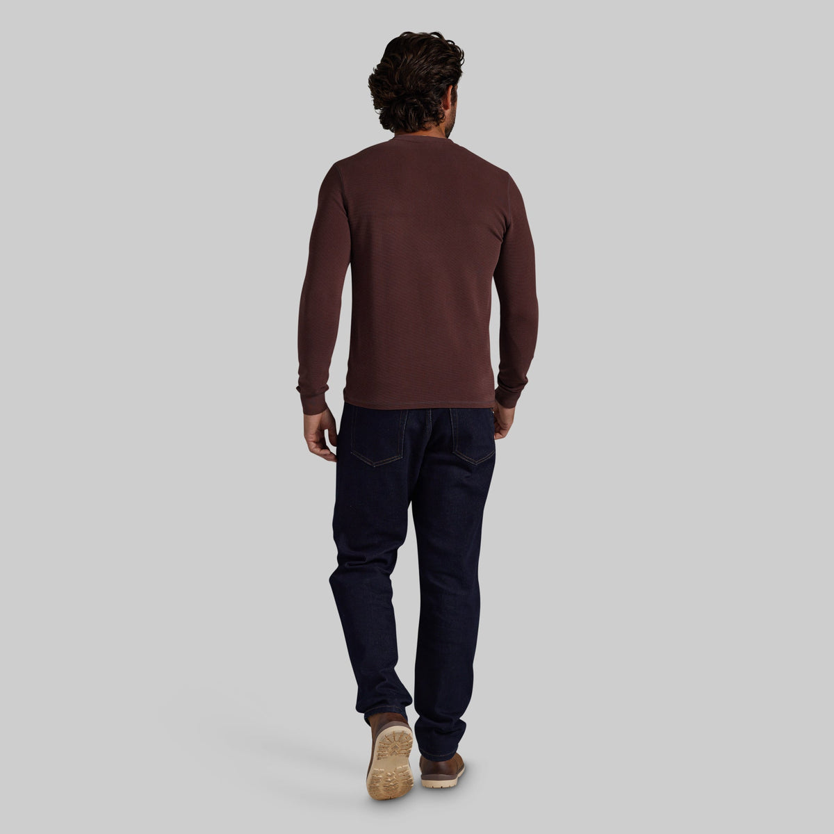A man viewed from behind wearing a dark brown long-sleeve crewneck shirt, dark navy jeans, and brown shoes, standing against a solid light gray background.
