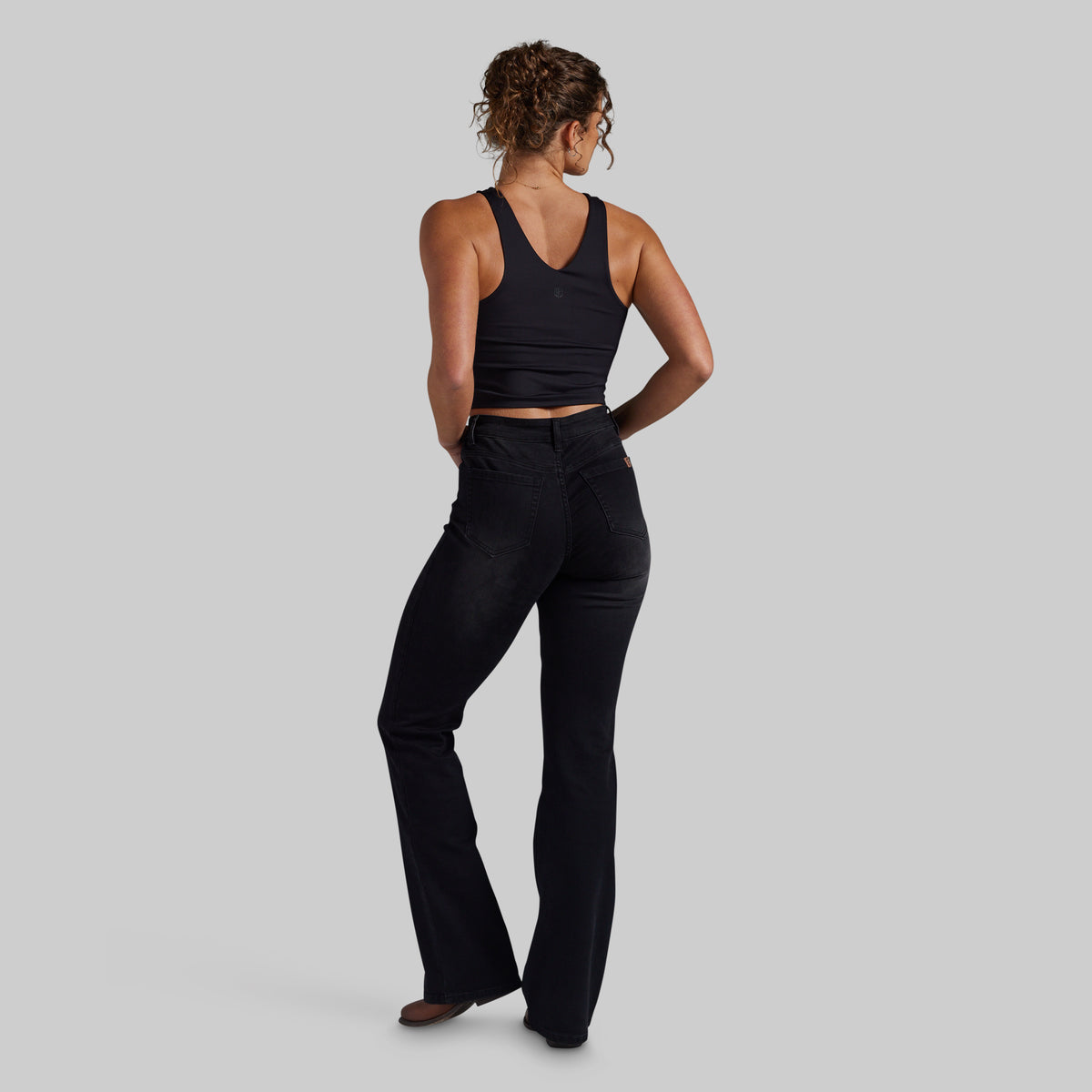 A woman is shown from the back wearing black mid-waisted bootcut jeans and a black ribbed crop top against a solid gray studio background.