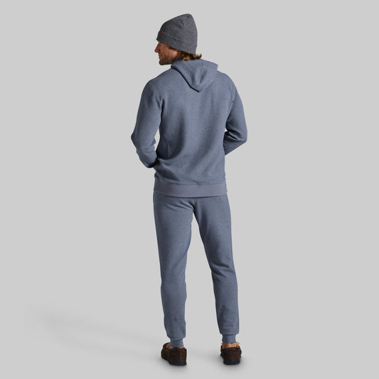 A man wearing a matching heathered slate blue hoodie and sweatpants set, viewed from behind, wearing a gray beanie and brown shoes, against a solid light gray background.