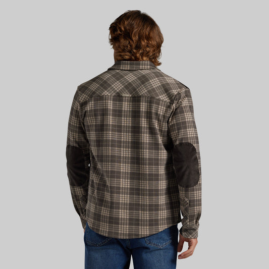 A man viewed from behind wearing a brown and beige plaid button-down shirt with dark brown elbow patches, paired with blue jeans, against a solid gray background.