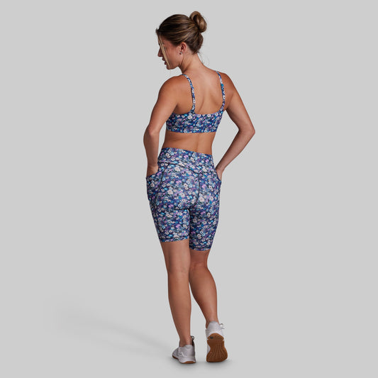 A woman is shown from the back wearing a matching athletic set consisting of a blue floral sports bra and biker shorts, set against a solid gray background.