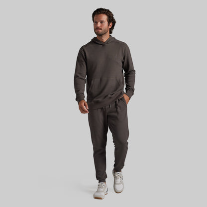A man modeling a dark grey matching hoodie and sweatpants set, paired with white sneakers, against a solid light gray background.