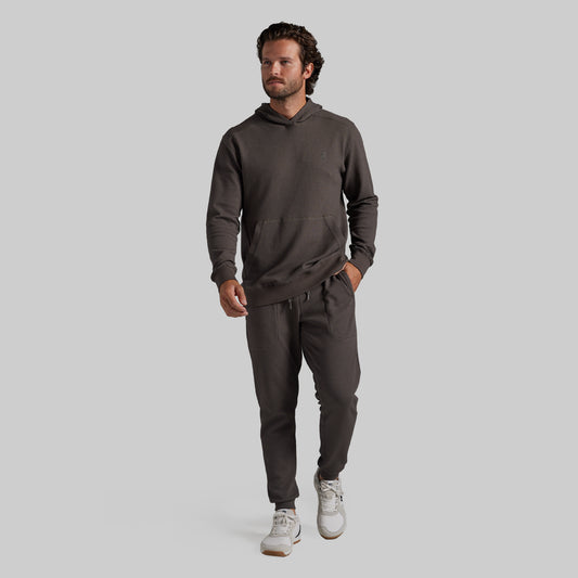 A man modeling a dark grey matching hoodie and sweatpants set, paired with white sneakers, against a solid light gray background.