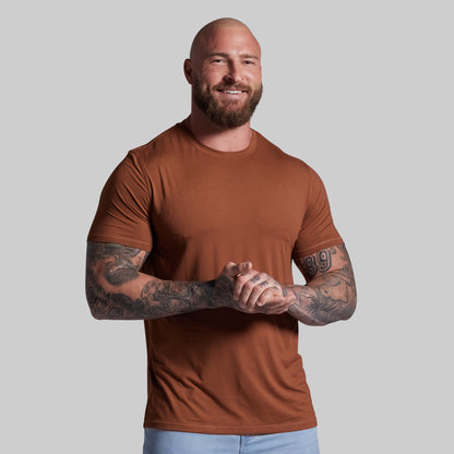 Man wearing a brown t-shirt with tattoos on his arms against a plain background