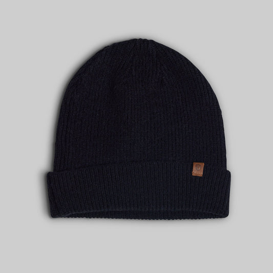 A dark navy blue ribbed knit beanie hat with a small brown leather brand patch on the cuff, shown against a solid gray background.