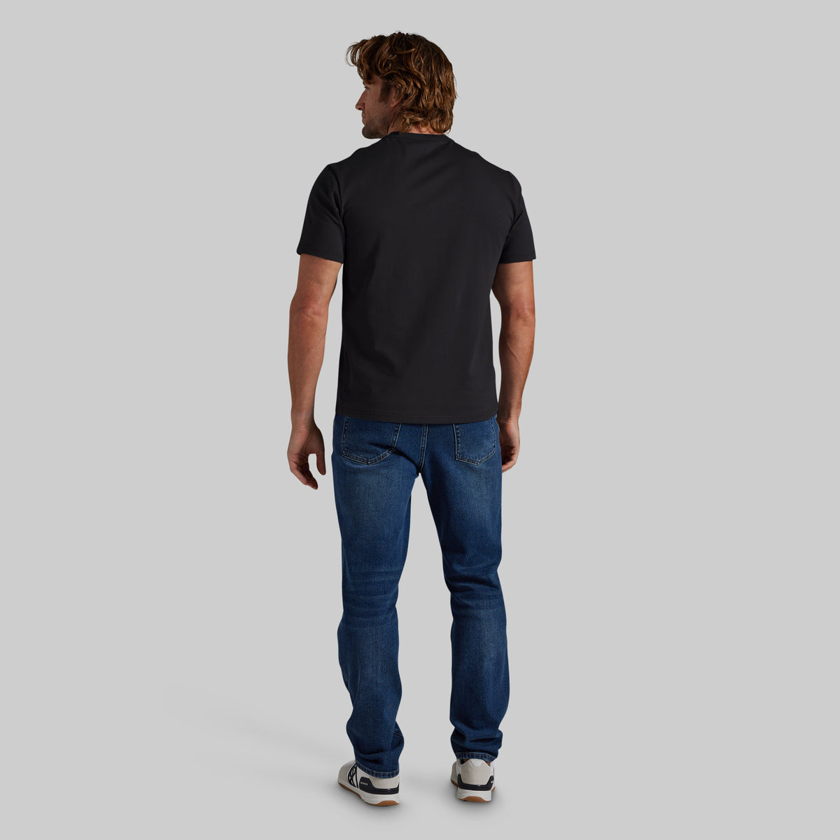 A man viewed from behind wearing a black short-sleeve t-shirt, blue jeans, and white sneakers, standing against a solid light gray background.