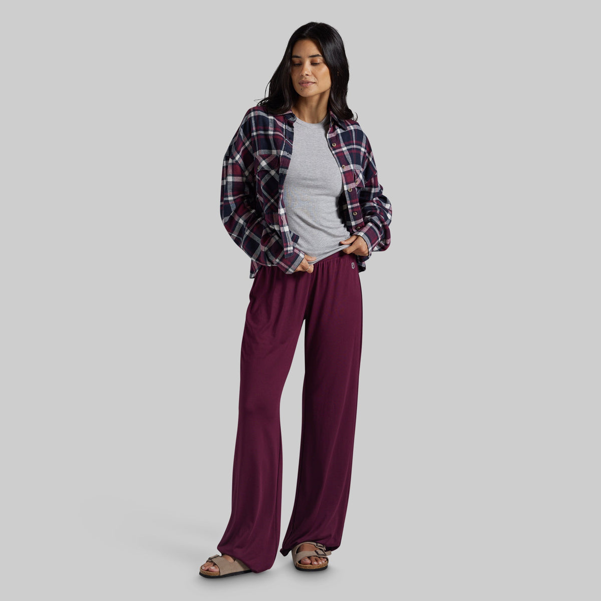 A woman modeling a pair of wide-leg burgundy pants, styled with a gray t-shirt, a dark red and navy plaid flannel shirt, and brown sandals, against a solid gray studio background.