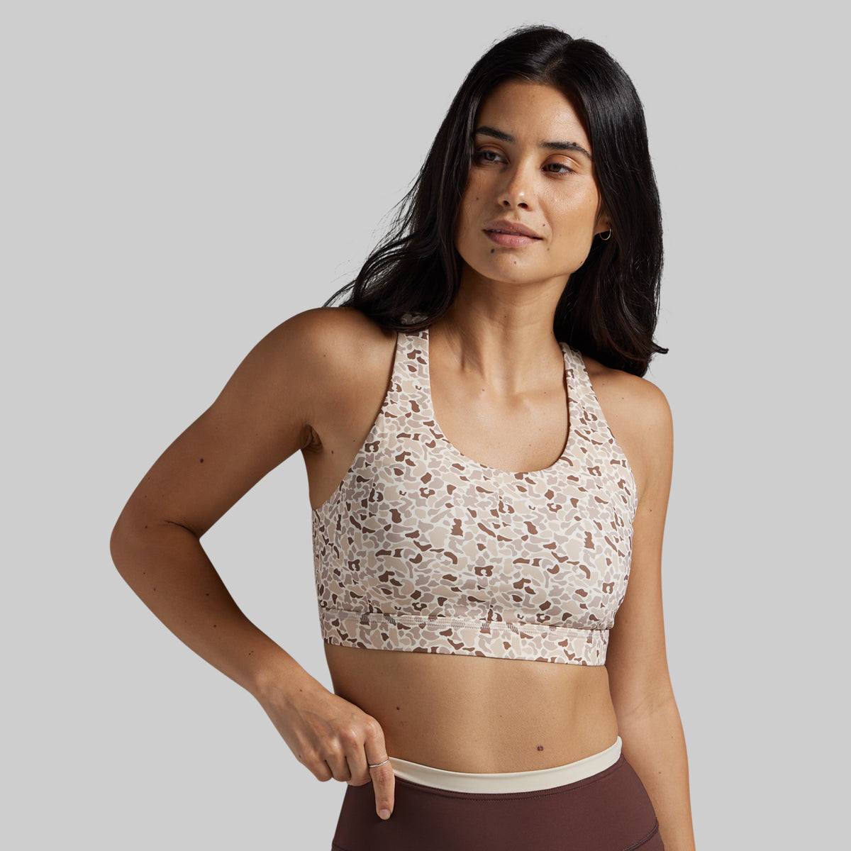 A woman modeling a beige sports bra with a brown abstract pattern, paired with brown leggings, against a solid gray background.