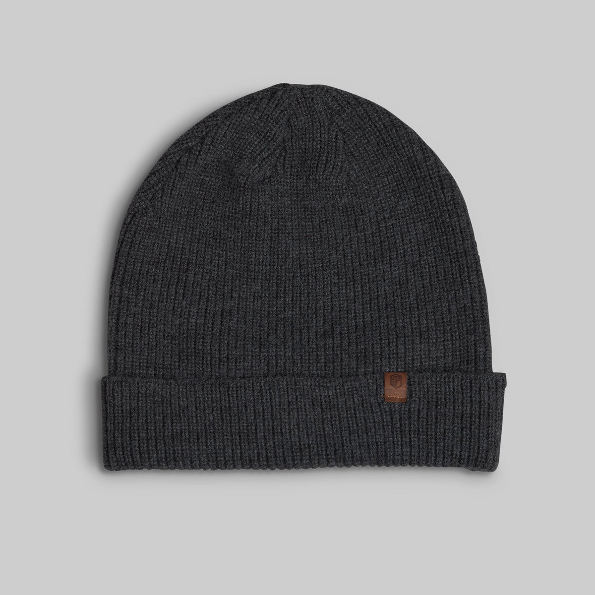 A dark charcoal gray ribbed knit beanie hat with a folded cuff and a small brown leather logo patch on the side, shown against a solid light gray background.