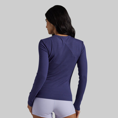 A woman shown from the back wearing a fitted long-sleeve athletic top in a deep purple shade, paired with light lavender shorts, against a solid light gray background.