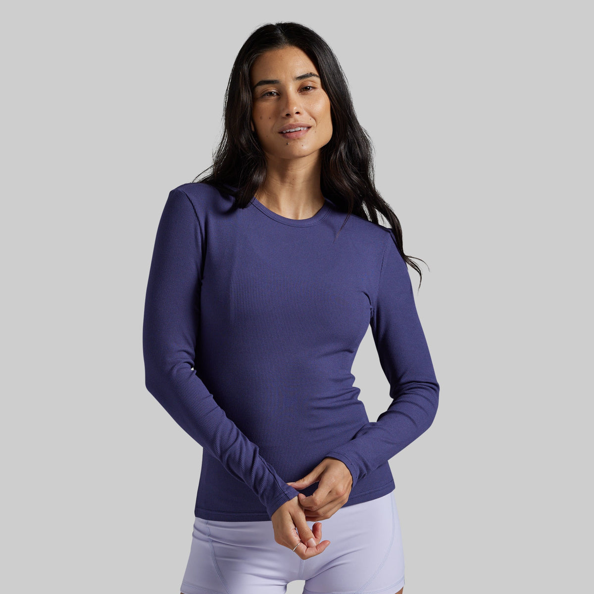 A woman modeling a long-sleeve crewneck athletic top in a solid periwinkle purple color, paired with white leggings, against a solid light gray background.