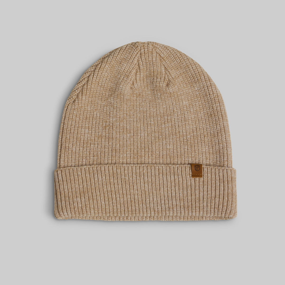 A beige ribbed knit beanie hat with a small brown leather brand patch on the folded cuff, shown against a solid gray background.