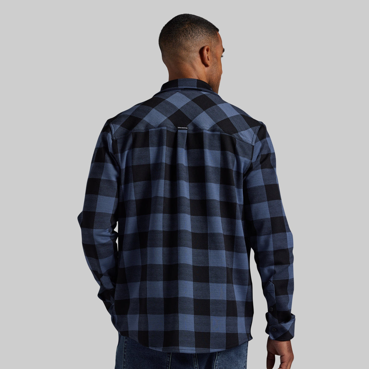 A man viewed from the back wearing a blue and black buffalo plaid flannel shirt, paired with blue jeans, against a solid gray background.