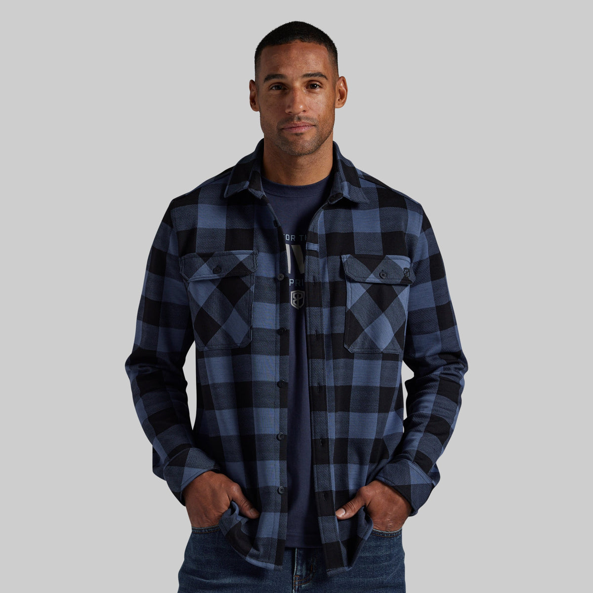 A man wearing a dark blue and black plaid button-up flannel shirt over a navy t-shirt, paired with blue jeans, against a solid gray background.
