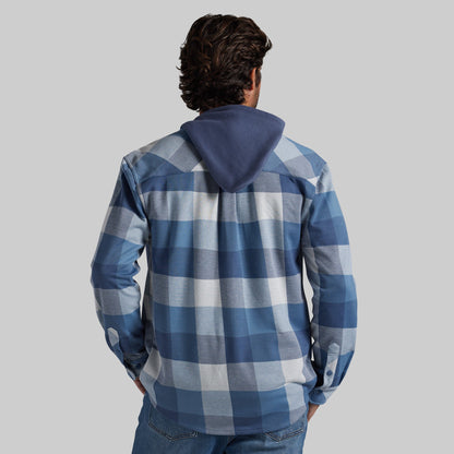 A man viewed from behind wearing a blue and white plaid hooded flannel with a solid navy blue hood, paired with blue jeans, against a solid gray background.