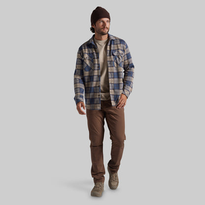 A man wearing a blue and beige plaid flannel shirt over a beige t-shirt, brown pants, and a dark brown beanie, walking against a solid gray background.