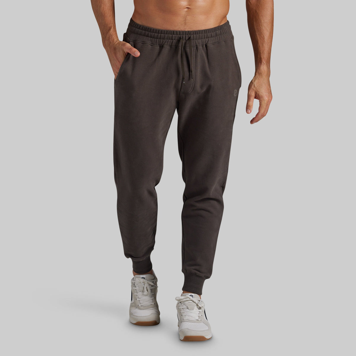 A man wearing dark grey jogger sweatpants with a drawstring waist and elastic cuffs, paired with white sneakers, shown from the waist down against a solid gray background.
