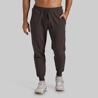 A man wearing dark brown jogger sweatpants with an elastic drawstring waist and ribbed ankle cuffs, paired with white sneakers, shown from the waist down against a solid gray background.