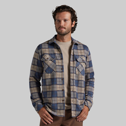 A man wearing a blue and beige plaid button-up flannel shirt over a beige t-shirt and brown pants, photographed against a solid gray studio background.