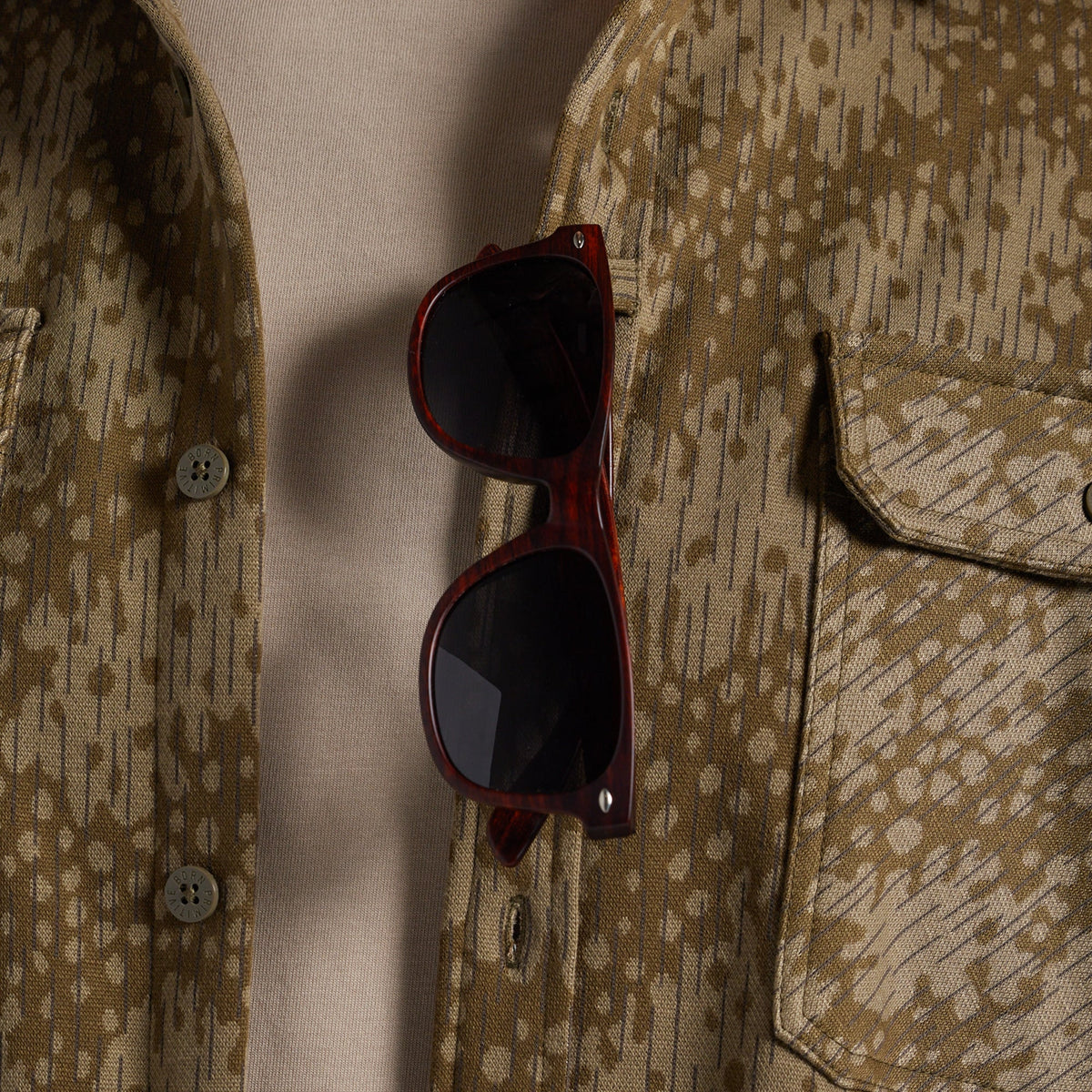 A close-up of a pair of dark brown wooden-framed sunglasses with black lenses, resting on the lapel of a patterned beige and brown button-up shirt.