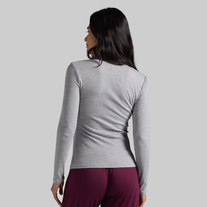 A woman shown from the back wearing a light heather gray long-sleeve athletic top and dark burgundy pants against a solid light gray background.