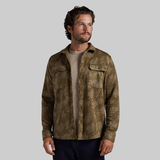 A man wearing a brown and olive green textured camo-print button-up shirt over a beige t-shirt and dark pants, photographed against a solid gray background.