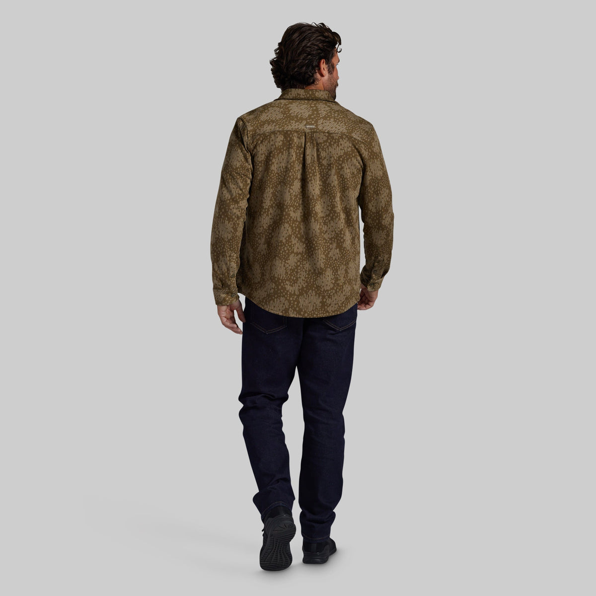 A man viewed from behind wearing a brown and olive patterned button-down shirt, dark navy pants, and black shoes, standing against a solid gray studio background.