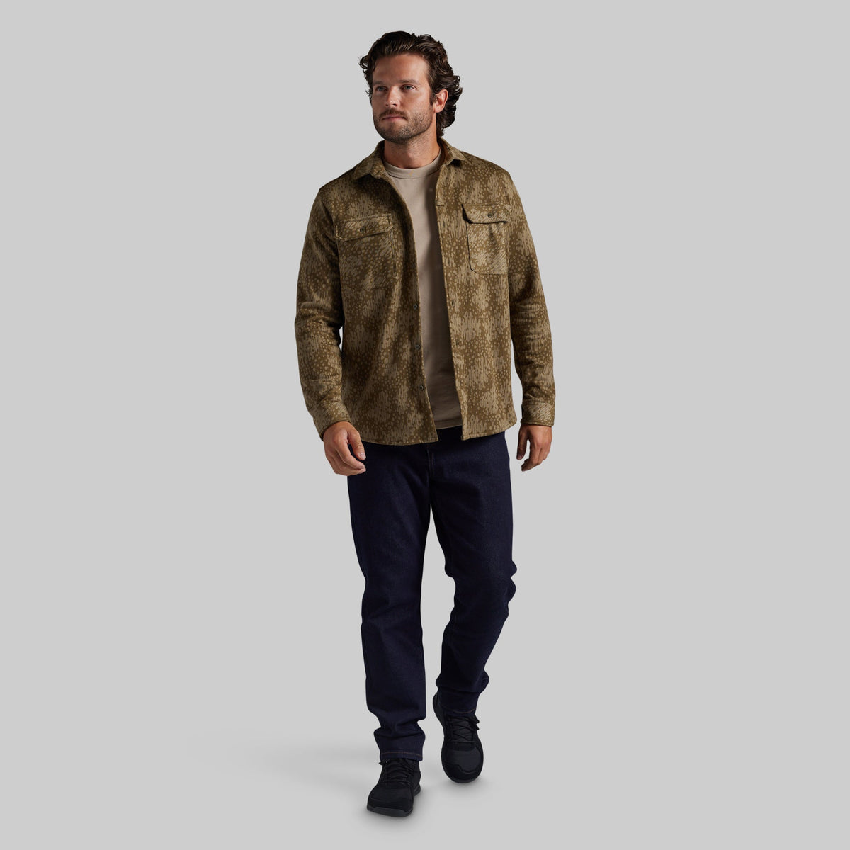 A man modeling a brown and beige textured jacket over a beige t-shirt, dark navy pants, and black boots, set against a solid gray background.