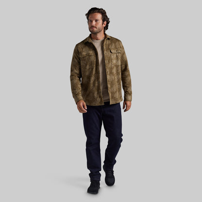 A man modeling a brown and beige textured jacket over a beige t-shirt, dark navy pants, and black boots, set against a solid gray background.