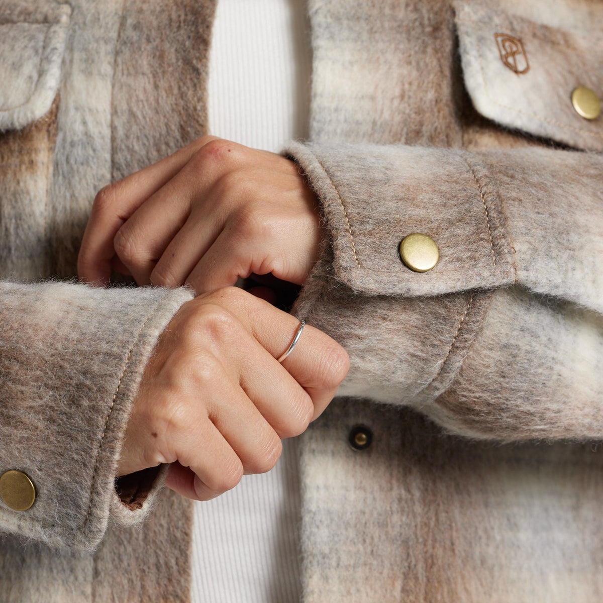 Close-up of a person wearing a soft, fuzzy plaid jacket in muted beige, taupe, and gray tones, with gold-tone snap buttons and a white ribbed top visible underneath.