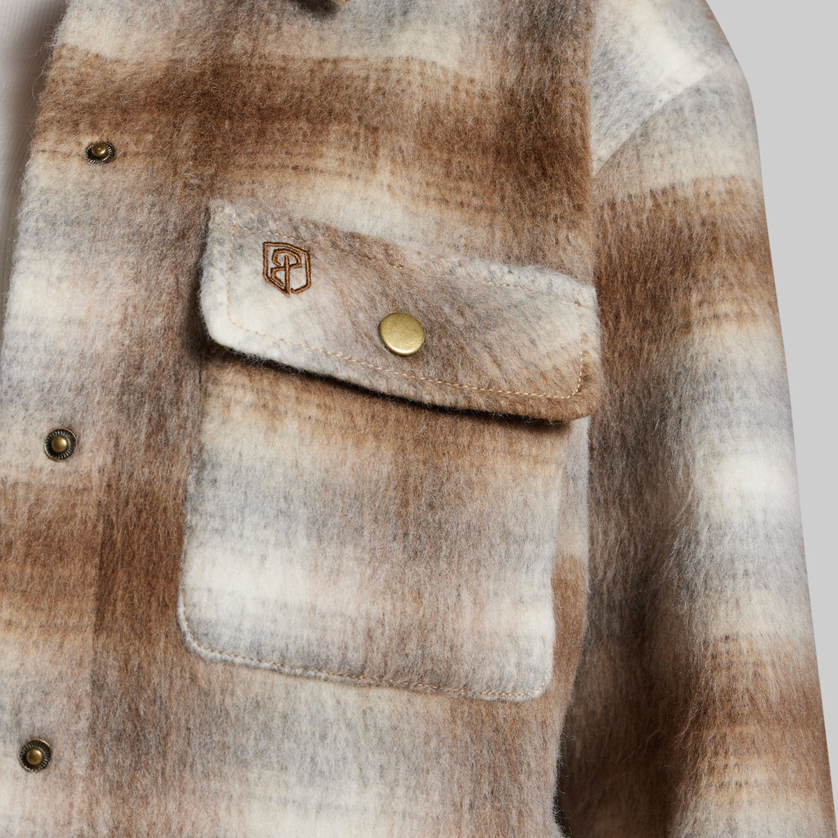 Close-up of a fuzzy plaid jacket in brown, beige, and cream tones, showing a chest pocket with a flap, brass-tone snap, and a small embroidered monogram logo.