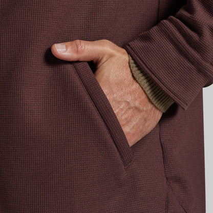 Close-up of a person's hand in a pocket of a textured brown waffle-knit jacket, with a ribbed olive green undershirt cuff visible.
