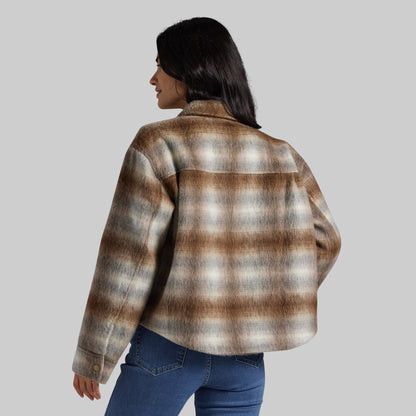 A woman shown from the back wearing a brown, beige, and gray plaid flannel shirt jacket with blue jeans, set against a solid gray studio background.
