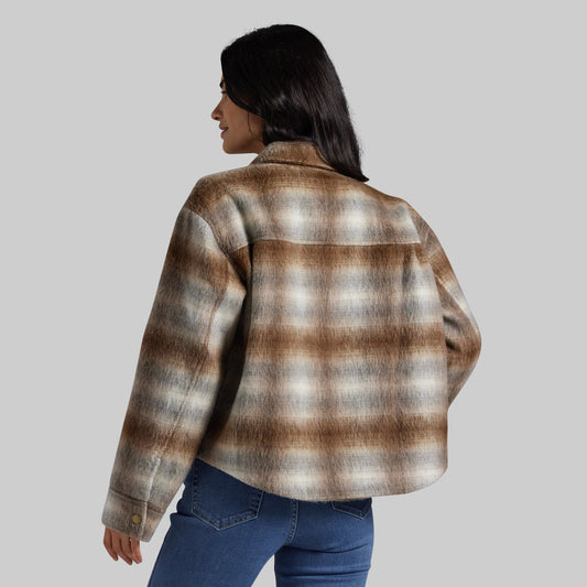 A woman shown from the back wearing a brown, beige, and gray plaid flannel shirt jacket with blue jeans, set against a solid gray studio background.