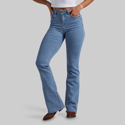 A person wearing light blue high-rise flare jeans with hands in pockets, paired with a white cropped top and brown pointed-toe heels, against a plain light gray studio background.