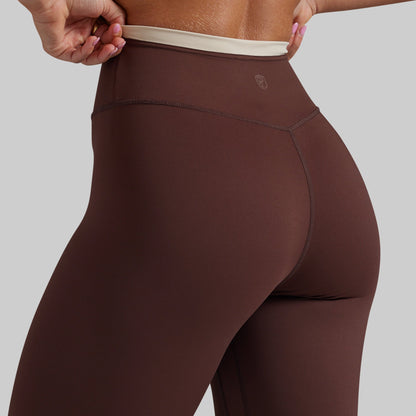 Close-up of a model wearing high-waisted brown leggings with a beige waistband, shown from the back side against a solid gray background.