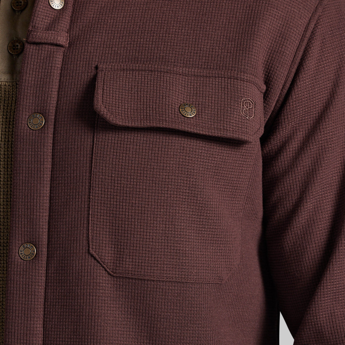 Close-up of a waffle-knit button-front shirt jacket in a muted burgundy-brown color, featuring a chest pocket with a flap and metal snap, plus a small embroidered logo.