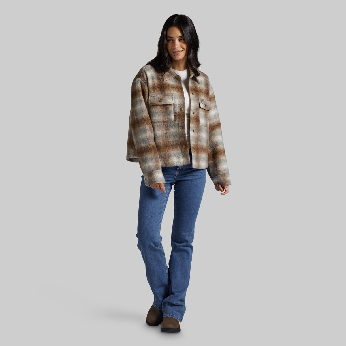A woman modeling a brown and beige plaid button-up flannel shirt jacket over a white t-shirt, paired with blue jeans and brown boots, against a solid gray background.