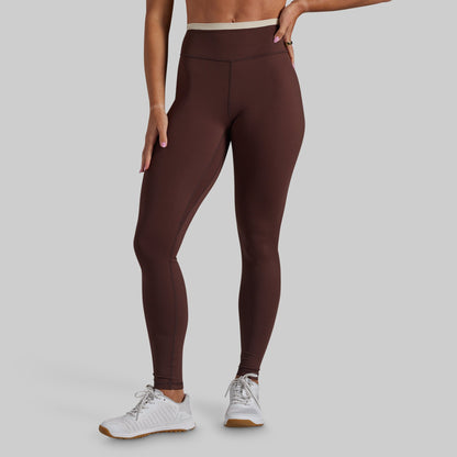 A model wearing dark brown high-waisted leggings and white athletic sneakers, shown from the waist down against a solid light gray background.