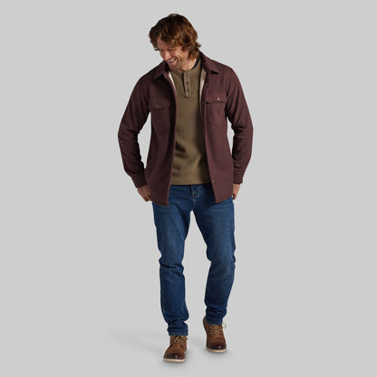 A man modeling a dark brown jacket over a brown henley sweater, paired with blue jeans and brown shoes, against a solid gray background.