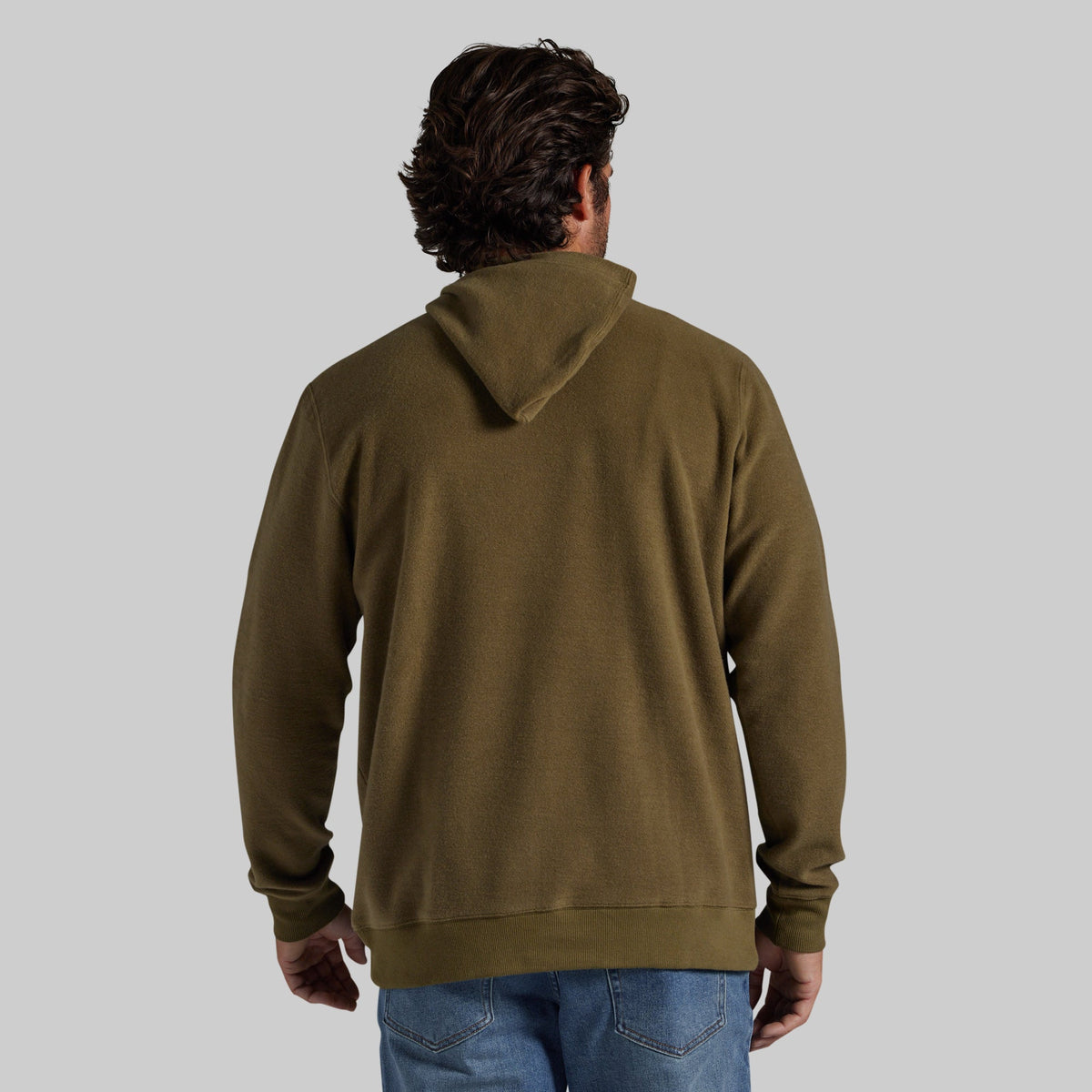 A man viewed from behind wearing an olive green pullover hoodie and blue jeans, standing against a solid light gray background.