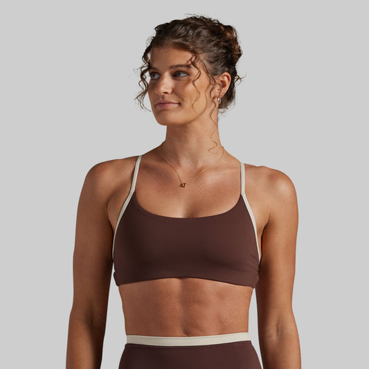 A woman wearing a brown sports bra with contrasting beige straps and a matching beige waistband, shown against a solid light gray background.