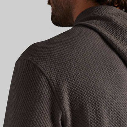Close-up of a man wearing a dark gray waffle-knit hooded sweatshirt, showing the textured fabric and collar from a side-back angle against a light gray background.