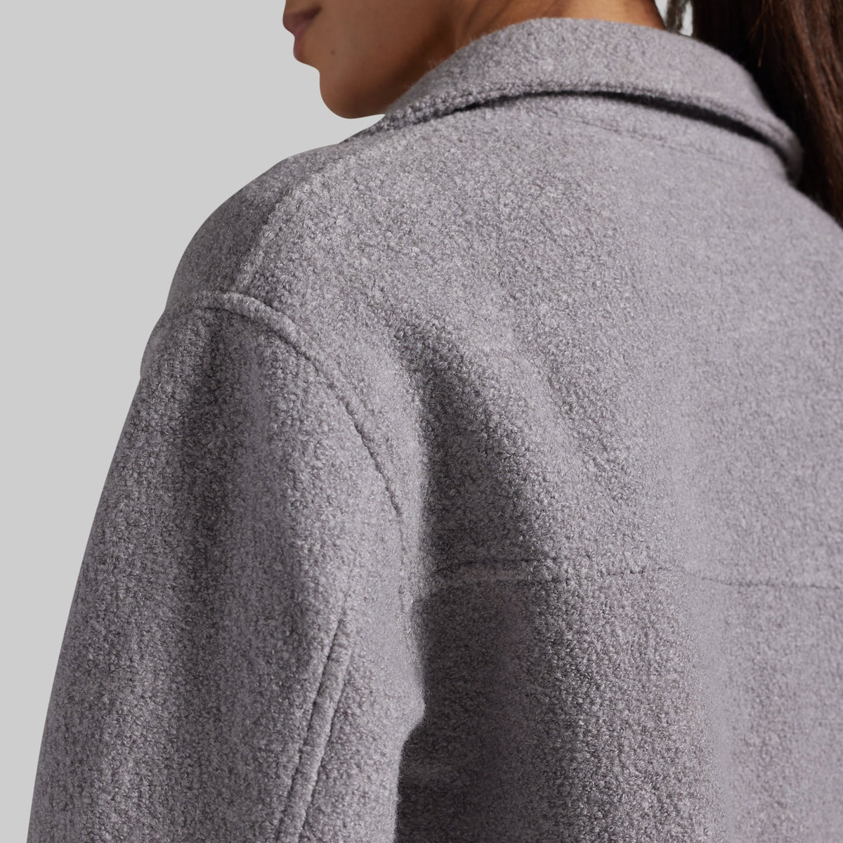 Close-up of a person wearing a textured light gray fleece or boucle jacket, showing the back and shoulder area against a plain light gray background.
