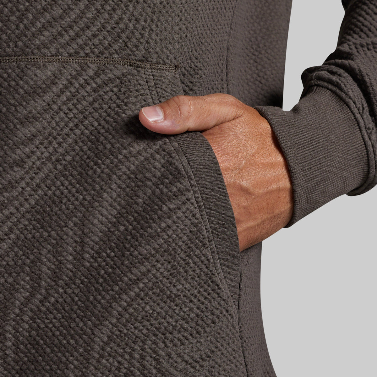 Close-up of a person wearing a dark grey textured pullover hoodie, showing a hand in the side pocket against a light gray background.
