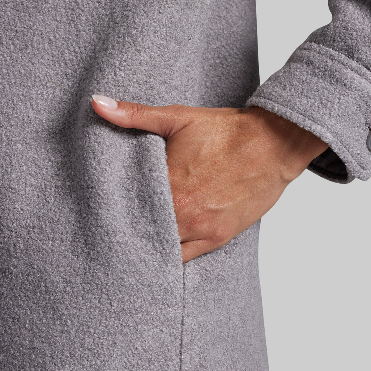 Close-up of a person wearing a heather gray textured coat, with a hand reaching into a side pocket against a light gray background.