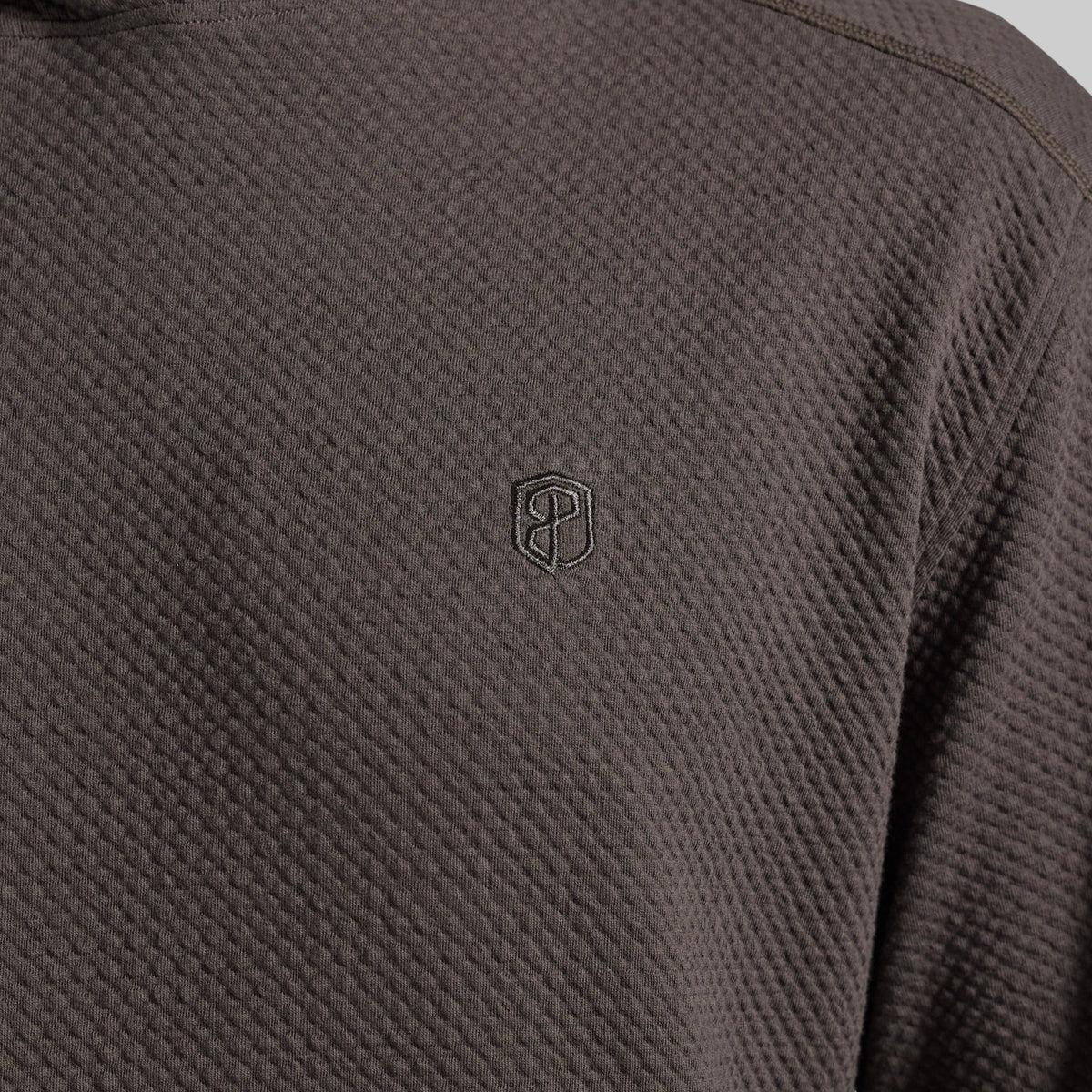 Close-up of a dark grey textured hoodie with a small tonal embroidered shield logo on the chest.