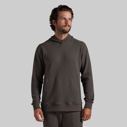A man wearing a dark brown/grey textured waffle-knit hoodie and matching sweatpants, standing against a solid light gray background.