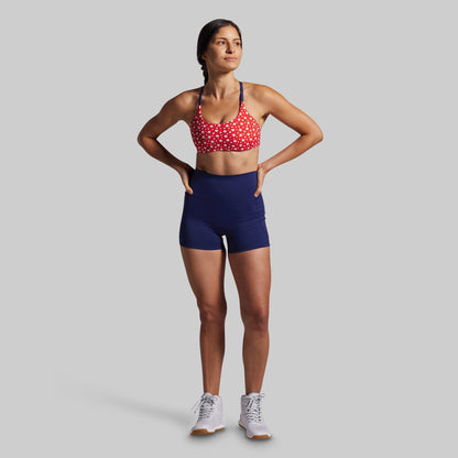 Woman wearing a red sports bra with white patterns and blue shorts on a gray background